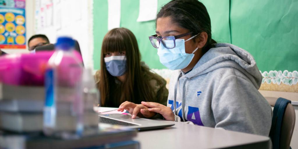 two girls look at a laptop in a classroom